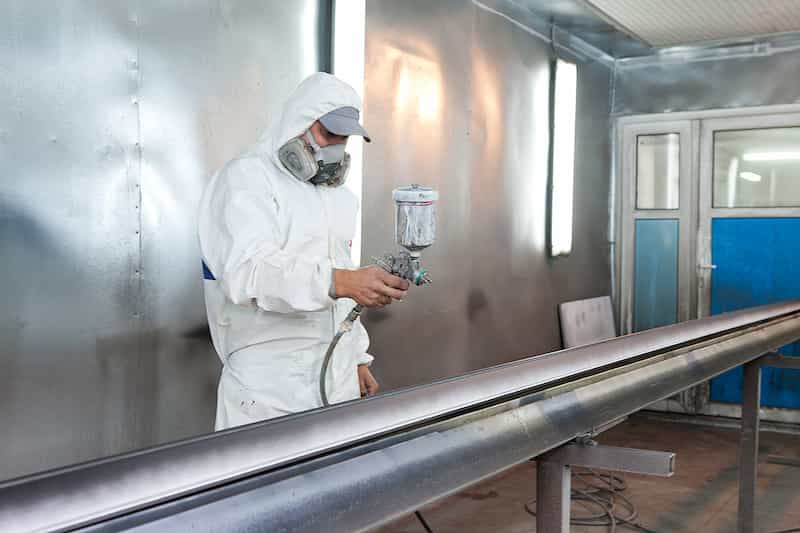 specjalista w kombinezonie ochronnym maluje profil okienny metodą natryskową w komorze lakierniczej